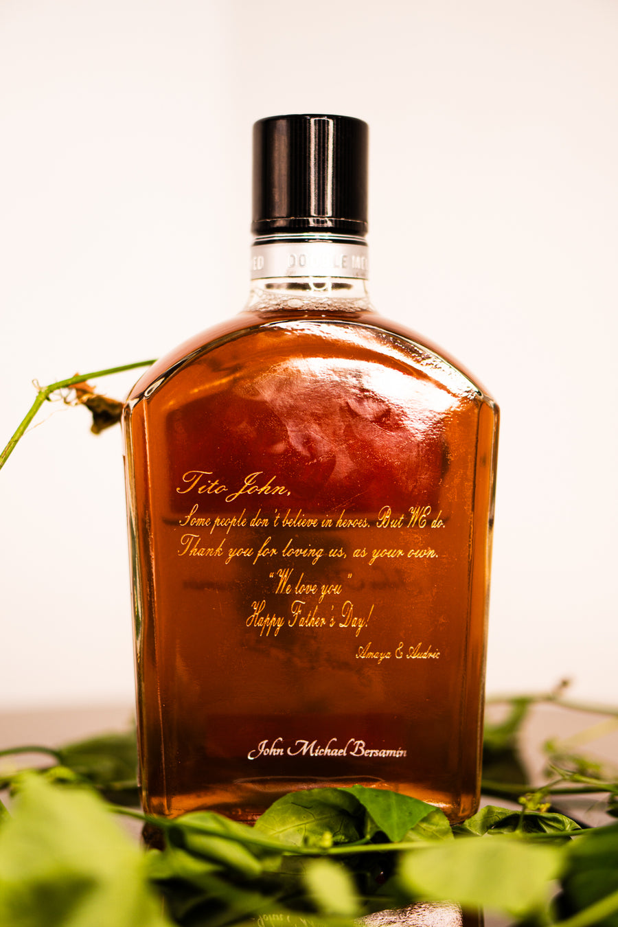 A custom engraved whiskey bottle with a black cap, filled with amber liquid, surrounded by green leaves on a white background.